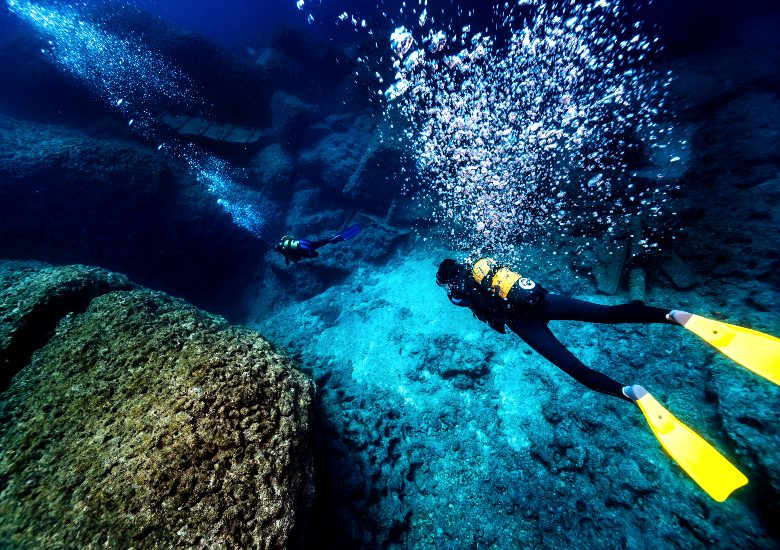 Scuba divers in clear Mediterranean water swimming towards a shipwreck